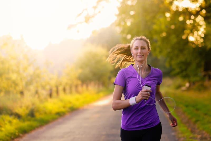 A person with long hair jogging on a path surrounded by trees during sunset. They are wearing a purple t-shirt, holding a smartphone, and have white earphones plugged into their ears. The path is lit by the warm glow of the setting sun.