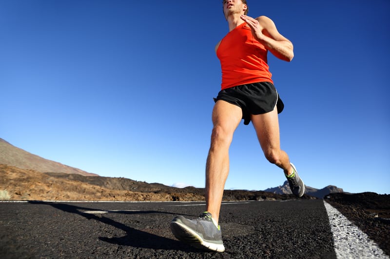 A person wearing a bright orange tank top, black shorts, and running shoes is jogging on an open road. The background features clear blue skies and barren, rocky terrain. The shot is taken from a low angle, capturing the person's legs mid-stride.