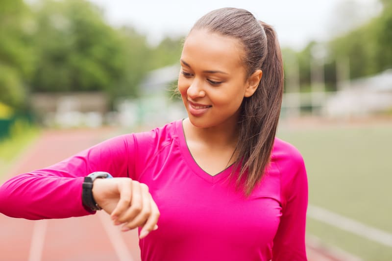 A woman in a bright pink long-sleeve shirt smiles while looking at her smartwatch. She is standing on an outdoor running track with a green field and trees in the background. Her hair is pulled back into a ponytail.