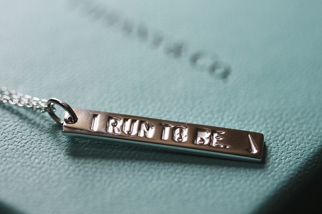 A silver bar-shaped pendant engraved with the words “I RUN TO BE.” is displayed against a blurred teal background, likely a branded Tiffany & Co. box.