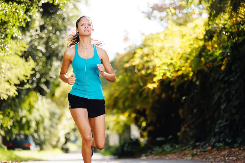 A person wearing a teal tank top and black shorts is jogging along a tree-lined path on a sunny day. They have earphones in, suggesting they might be listening to music while exercising. The background is lush with green foliage.