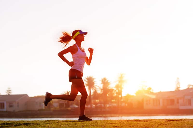 A woman in athletic clothing, including a tank top, shorts, and a visor, is running outdoors at sunset. The scene includes grassy terrain and houses in the background, with the sun low on the horizon, casting a warm golden light.