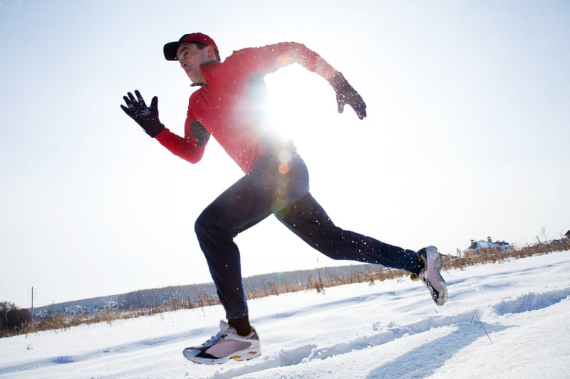 A person wearing a red jacket, black pants, gloves, and a cap is running energetically through snow-covered ground. The sun shines brightly behind, creating a radiant effect and casting long shadows on the snow.