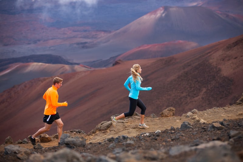 Two people are trail running on a rugged path surrounded by a volcanic landscape. The person in the foreground wears a teal long-sleeve top and black leggings, while the person behind wears a bright orange long-sleeve top and black shorts.