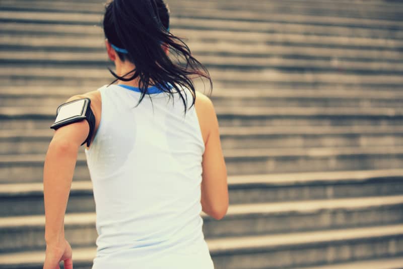 A person with long hair, wearing a white tank top and a fitness armband, is running up a series of outdoor steps. The background is a blurred view of the steps, emphasizing the upward motion and effort.