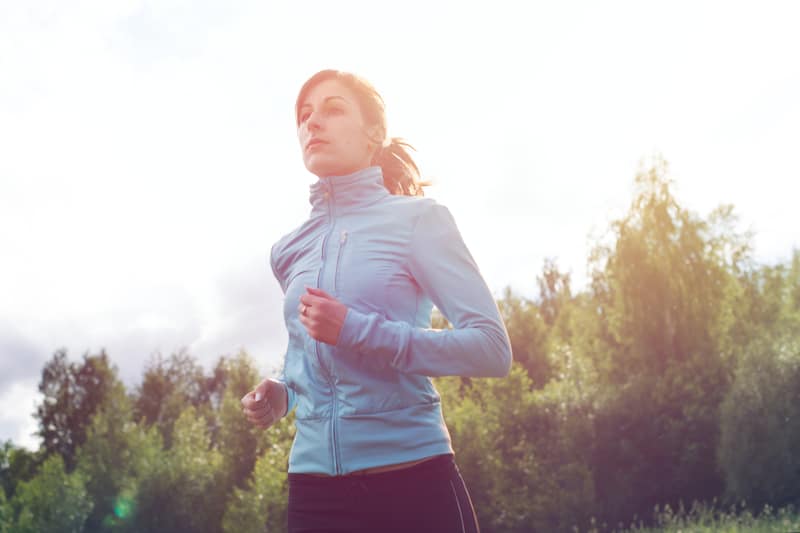 A person jogs outdoors under a bright sky. They are wearing a light blue jacket and dark pants. Trees in the background are slightly blurred, suggesting motion. The sun shines brightly, giving the scene a serene and energetic atmosphere.