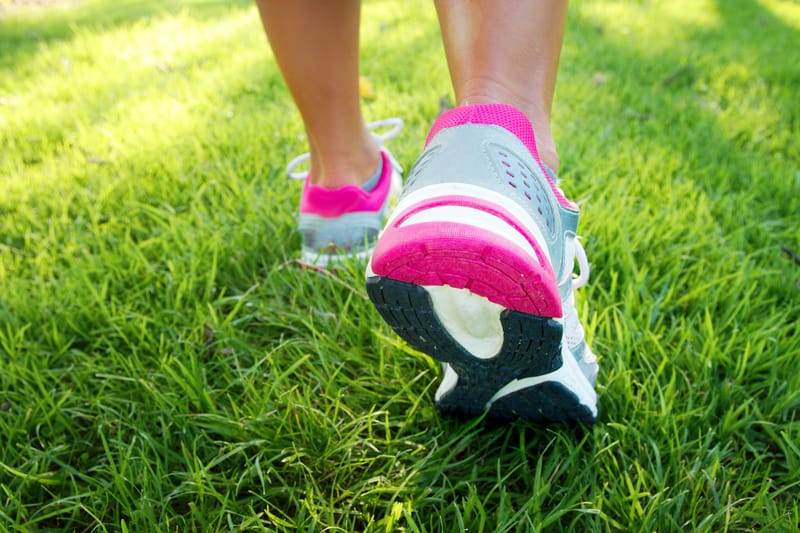 Close-up of a person walking on a grassy field, focusing on their lower legs and sports shoes. The shoes are grey with pink accents and black soles, while the vibrant green grass suggests a sunny, outdoor setting.