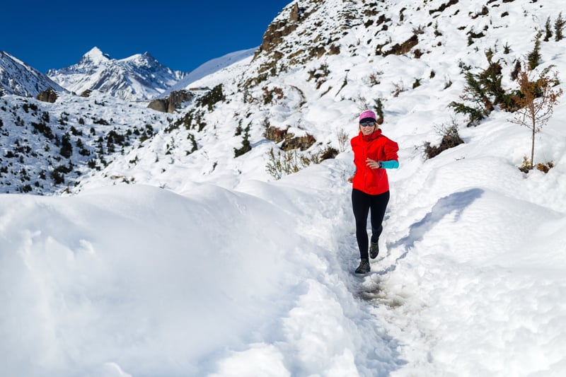 A person dressed in a red jacket and black pants runs through a narrow, snowy path in a mountainous landscape. The sun is shining brightly on the snow-covered mountains and trees, and the sky is clear and blue.