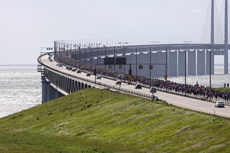 A large crowd of people walking on a bridge over water, with several vehicles on the road alongside them. The bridge curves gently and disappears into the distance. The foreground includes a grassy slope with wildflowers. The sky is clear and sunny.