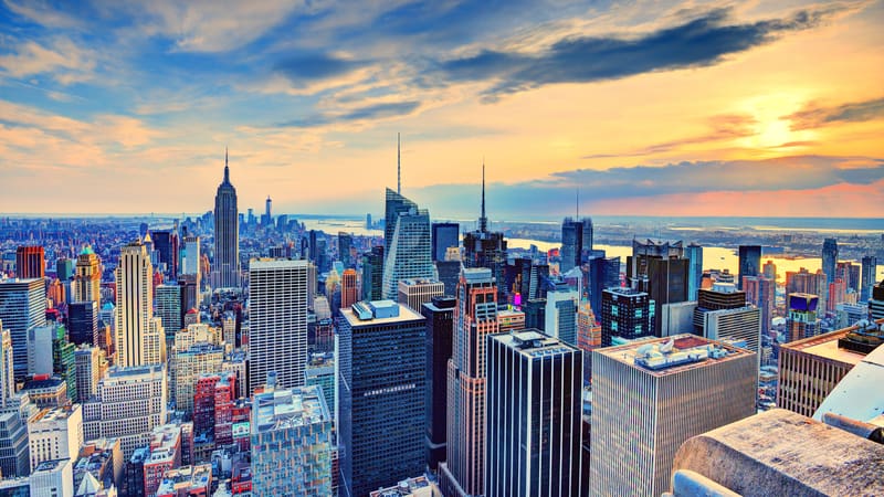 Aerial view of a vibrant cityscape at sunset. The skyline is adorned with tall skyscrapers including iconic structures. The sky is a gradient of yellows, oranges, and blues, as the sun descends, casting a warm glow on the buildings.