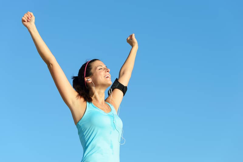 A woman in a light blue tank top is outdoors, raising her arms in triumph against a clear blue sky. She is smiling, wearing headphones, and has a fitness armband on her upper arm.