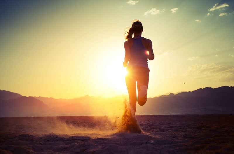 A person runs on a sandy terrain at sunset, creating a dust trail behind. The sun is low, casting a warm glow and long shadows, with mountains in the background. The sky is clear with some scattered clouds.