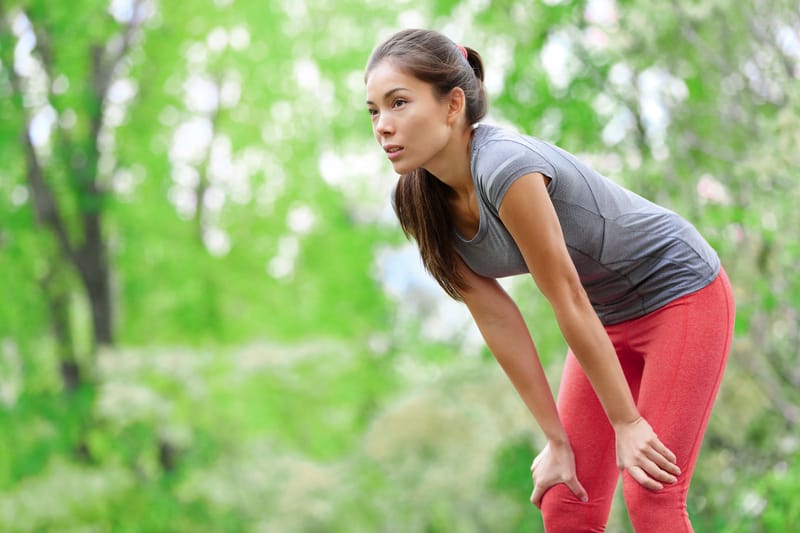 A woman dressed in a gray t-shirt and red pants is taking a break outdoors. She leans forward with her hands on her knees, looking focused. The background is lush and green, indicating she is in a park or a natural setting.