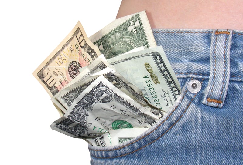 A close-up image of a person's back pocket of blue jeans filled with U.S. dollar bills, including $1, $5, $10, and $20 denominations. The pocket is partially open, showing the money stuffed inside. The background is white.