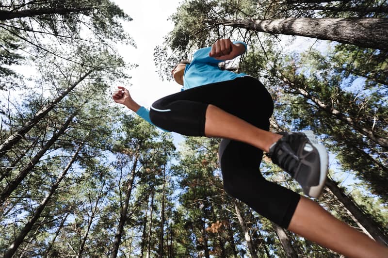 A person in athletic attire is running outdoors, captured from a low angle. They are wearing a blue shirt, black leggings, and gray sneakers. Large trees and a blue sky are visible above, creating a refreshing forest environment.