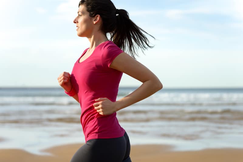 A woman with dark hair in a ponytail is jogging along a sandy beach. She is wearing a pink T-shirt and dark leggings. The ocean and a clear sky are in the background.