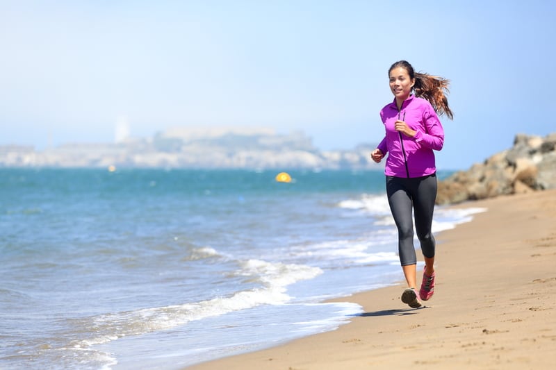 A woman in a pink jacket and black leggings runs along a sandy beach with gentle waves. The ocean is calm, and there are blurred buildings or structures visible on the horizon in the background. The sky is clear and blue.