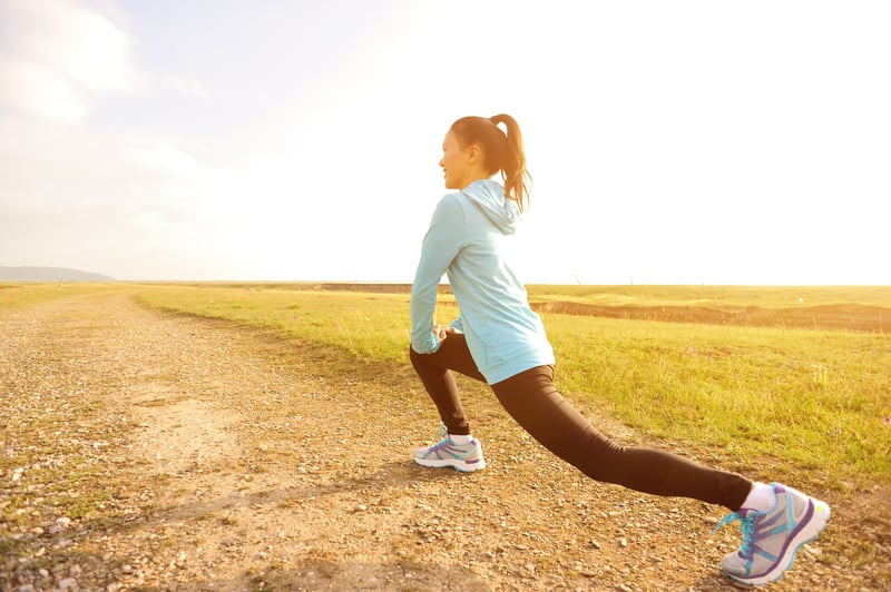 A woman in athletic clothing stretches by lunging forward on a gravel path in a rural area. She is wearing a light blue hoodie, black leggings, and running shoes. The sun is shining brightly in the background, casting a warm glow over the scene.