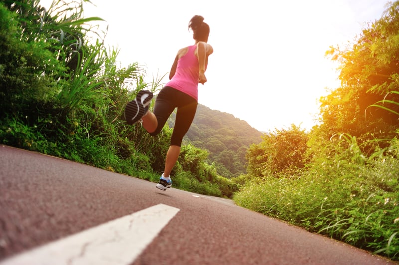 A woman in a pink tank top and black leggings runs on a paved road lined with greenery. The sun is shining brightly in the background, casting a warm glow. She is captured mid-stride, moving away from the camera.