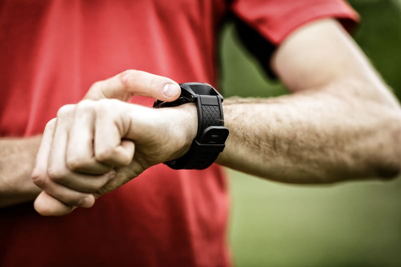 A person in a red shirt is checking their smartwatch. One finger is touching the screen of the watch, and the background is blurred green.