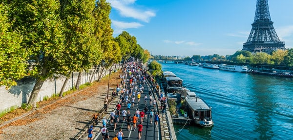 A large group of marathon runners jogs along a scenic riverside pathway framed by lush green trees on one side and the river on the other, with the iconic Eiffel Tower standing tall in the background under a bright blue sky.