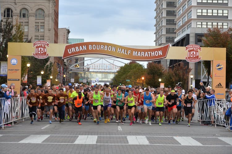 Runners at the starting line of the Urban Bourbon Half Marathon on a city street, enclosed by cheering spectators behind barricades. The starting banner is elevated, displaying the race name. Buildings and trees with autumn foliage line the background.