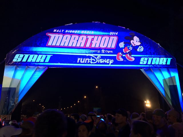 A large crowd is gathered under an illuminated arch at night, signaling the starting line for the Walt Disney World 2014 Marathon. The arch is decorated with an image of Mickey Mouse running and features both the Cigna and runDisney logos.