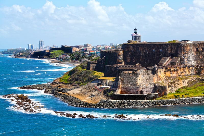 Historic fortifications of El Morro fortress stand prominently on the coast of San Juan, Puerto Rico, with rugged cliffs meeting the Atlantic Ocean's blue waves. The lighthouse atop the fort is visible against a backdrop of the cityscape and a partly cloudy sky.