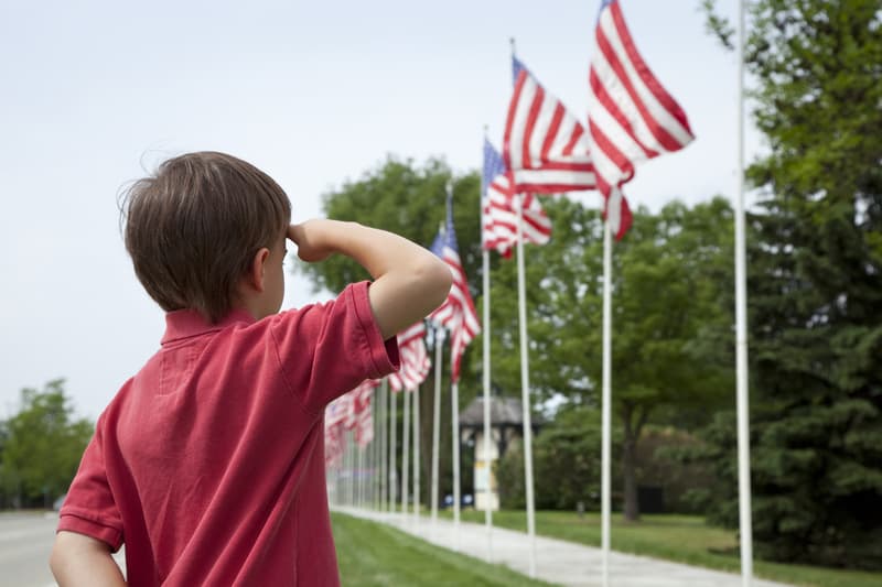 A young boy in a red shirt stands outdoors on a grassy area, saluting a row of American flags mounted on poles that line a paved path. Trees and bushes are visible in the background on a clear day.