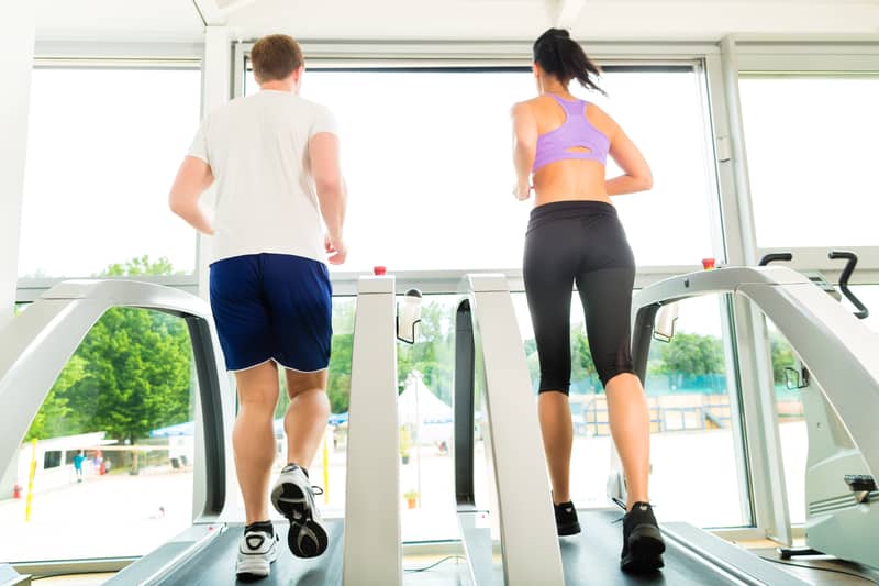 A man and a woman are running on side-by-side treadmills in a gym. Both are dressed in athletic wear; the man in a white t-shirt and blue shorts, and the woman in a purple sports bra and black leggings. Large windows in front of them reveal a bright outdoor scene.