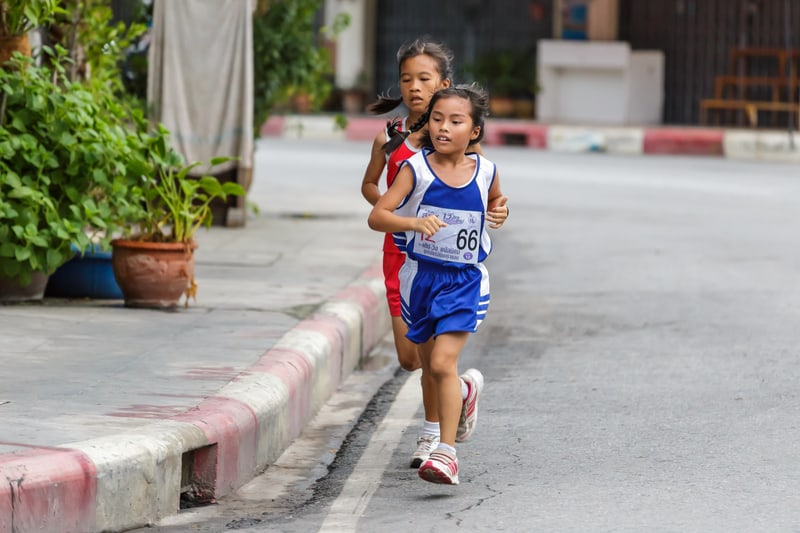 Two young girls are running in a race on a paved street. One girl, wearing a blue outfit with the number 66, is in the lead, while the other girl in a red outfit follows closely behind. Potted plants and a curb line the left side of the path. The background shows urban elements.