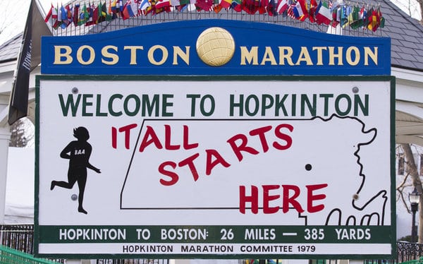 A decorative sign marks the start of the Boston Marathon in Hopkinton, featuring a map outline of Massachusetts, colorful international flags, a running figure, and text that reads: "Welcome to Hopkinton. It all starts here. Hopkinton to Boston: 26 miles, 385 yards.