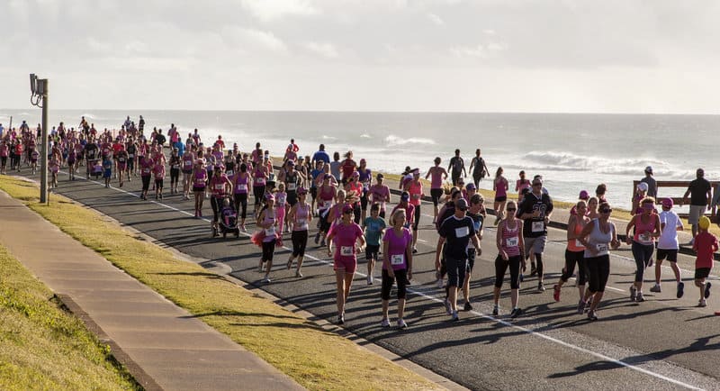 A large group of people participating in a marathon or running event by the sea. Many are dressed in pink, and the scene is set beside a coastal road with the ocean visible in the background. The weather is clear with a mix of sun and cloud.