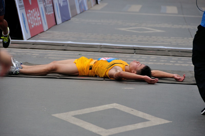 Runner in a yellow outfit lies on the pavement with arms spread out after finishing a race. The scene appears to be at the finish line, and the runner looks physically exhausted.