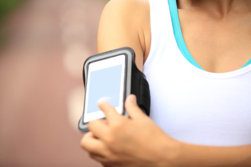 A person wearing a white tank top and a blue sports bra is adjusting a smartphone held in a black armband on their upper arm. The background is blurred and appears to be outdoors.