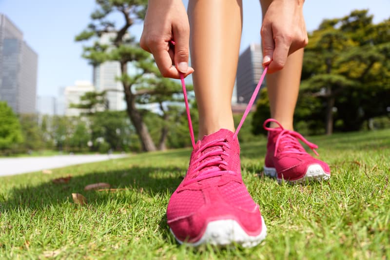 Close-up of a person outdoors, tying the laces on bright pink athletic shoes. The person is standing on lush green grass with leafy trees and tall buildings visible in the background on a sunny day.