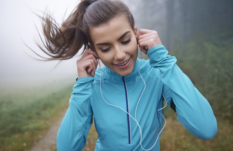 A woman in a blue jacket is jogging on a foggy trail while listening to music with white earphones. Her hair is tied back in a ponytail, and she is looking down, smiling. The background shows greenery and mist.