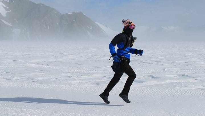 A person dressed in warm, blue and black winter gear runs across a snow-covered landscape with mountains in the background. They are wearing a hat, goggles, and facemask, and the sky is slightly overcast.