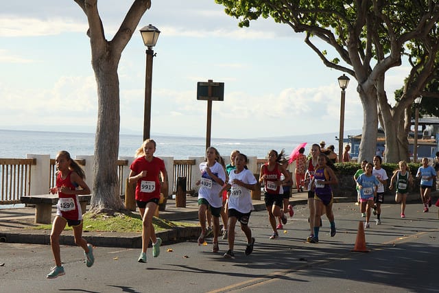 A group of runners participating in a race move along a scenic coastal road lined with trees. They wear colorful athletic clothing and numbered bibs. The sky is clear and the ocean is visible in the background. Spectators and street lamps are also present.