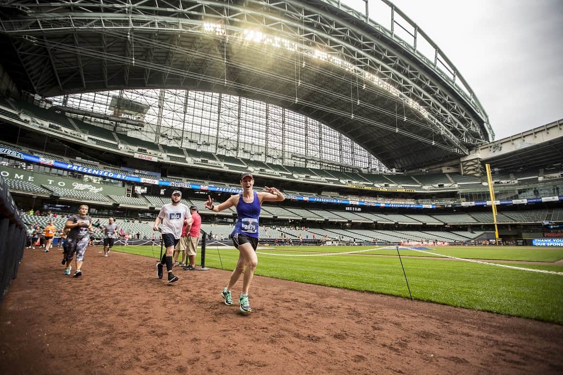 Participants in a race are running along the dirt path inside a large stadium with a retractable roof. The foreground shows a woman in athletic wear and a visor, while others follow behind. The stands are mostly empty, and the sky is partly cloudy.