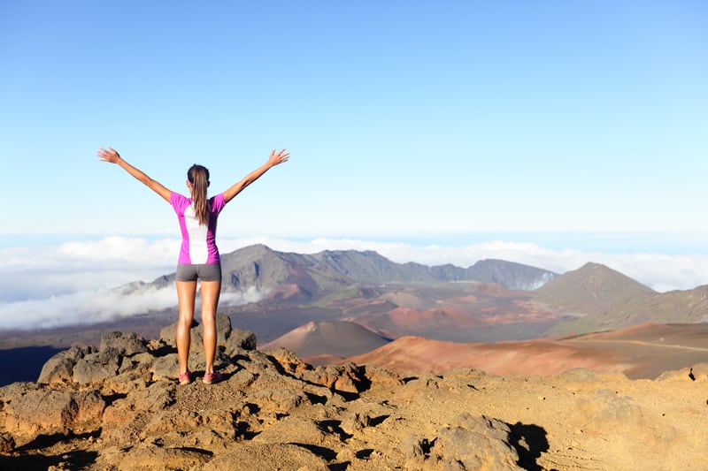 A person wearing a pink top and shorts stands on rocky terrain with arms raised, facing a distant mountain range under a clear blue sky. The person is above cloud level, overlooking a vast landscape of rugged mountains and valleys.