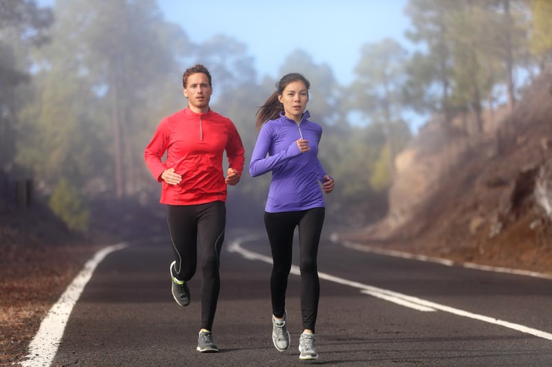 A man and woman jog on a paved road surrounded by trees on a foggy day. The man wears a red long-sleeve shirt and black pants, while the woman wears a purple long-sleeve shirt and black pants. Both are in motion, with trees and mist in the background.