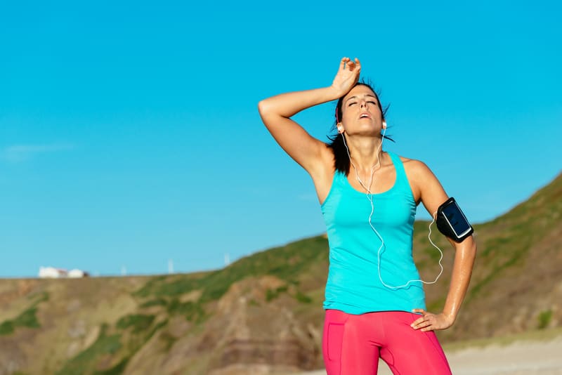 A woman wearing a turquoise tank top and pink leggings, with a phone armband and earphones, is taking a break from her run. She stands with one hand on her hip and the other on her forehead, appearing exhausted under a clear blue sky on a beach with hills in the background.