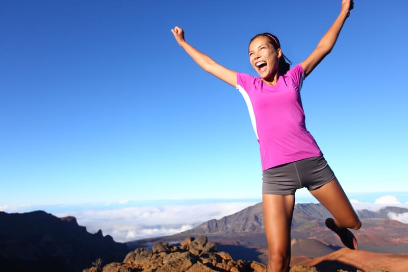 A woman in a pink shirt and gray shorts is jumping joyfully with her arms raised above her head. She is outdoors on a rocky landscape with mountains in the background under a clear blue sky.