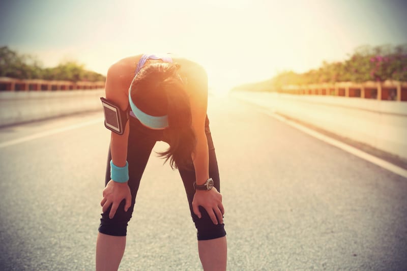 A person wearing blue fitness gear and a headband stands on an empty road, bent over with hands on knees, appearing exhausted under the bright sun. An armband with a phone is visible on their upper arm.