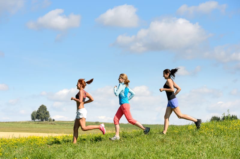 Three women jog in single file on a grassy field under a blue sky dotted with clouds. They wear athletic clothing with the woman in front in a white tank top and shorts, the second in blue and red attire, and the third in a black tank top and blue shorts.