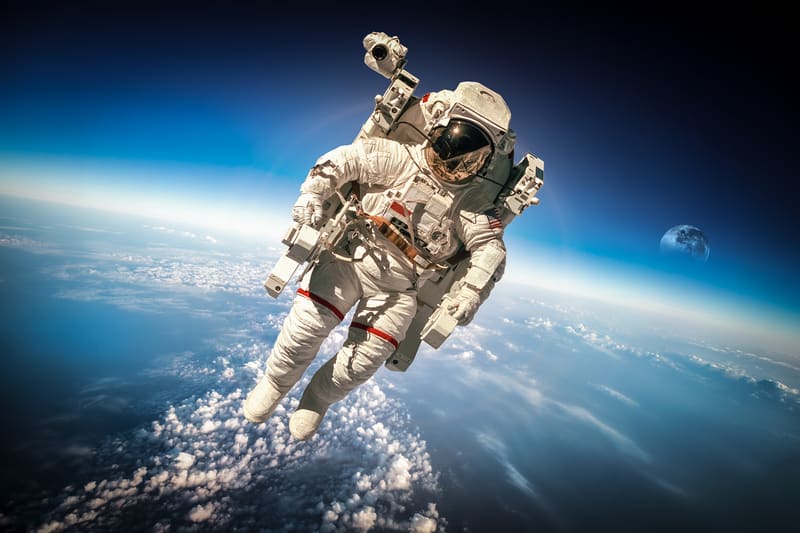 Astronaut in a spacesuit floats above Earth's atmosphere with clouds and the curvature of Earth visible below. The moon appears in the distant background against the dark expanse of space. The astronaut is performing a spacewalk, tethered by equipment.