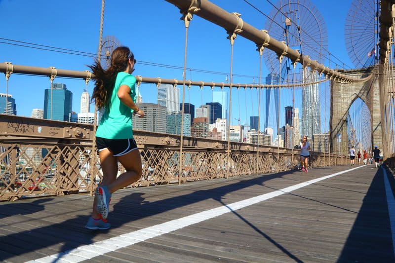 A woman in a green shirt and black shorts is jogging on a sunny day across a bridge with a city skyline in the background. Other people are walking and running on the bridge as well.