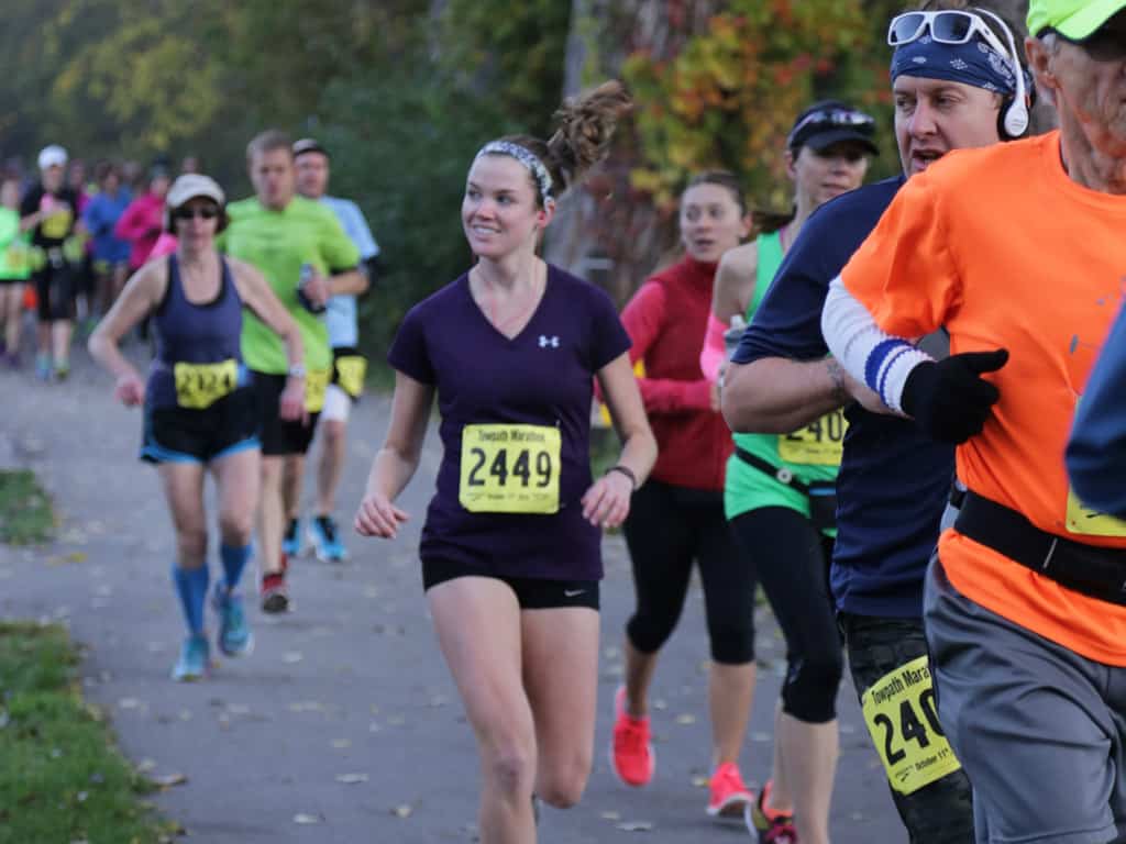 A group of runners participating in a marathon on a paved pathway. The runners are wearing various outfits, bib numbers, and some have headbands or hats. The background features trees, indicating a park-like setting. One runner in a purple shirt smiles while running.