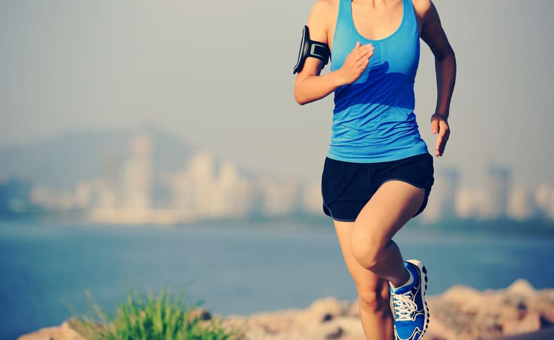 A person in athletic attire, including a blue tank top and black shorts, is running along a rocky shoreline. They have an armband device on their left arm. A blurred city skyline and water are visible in the background.